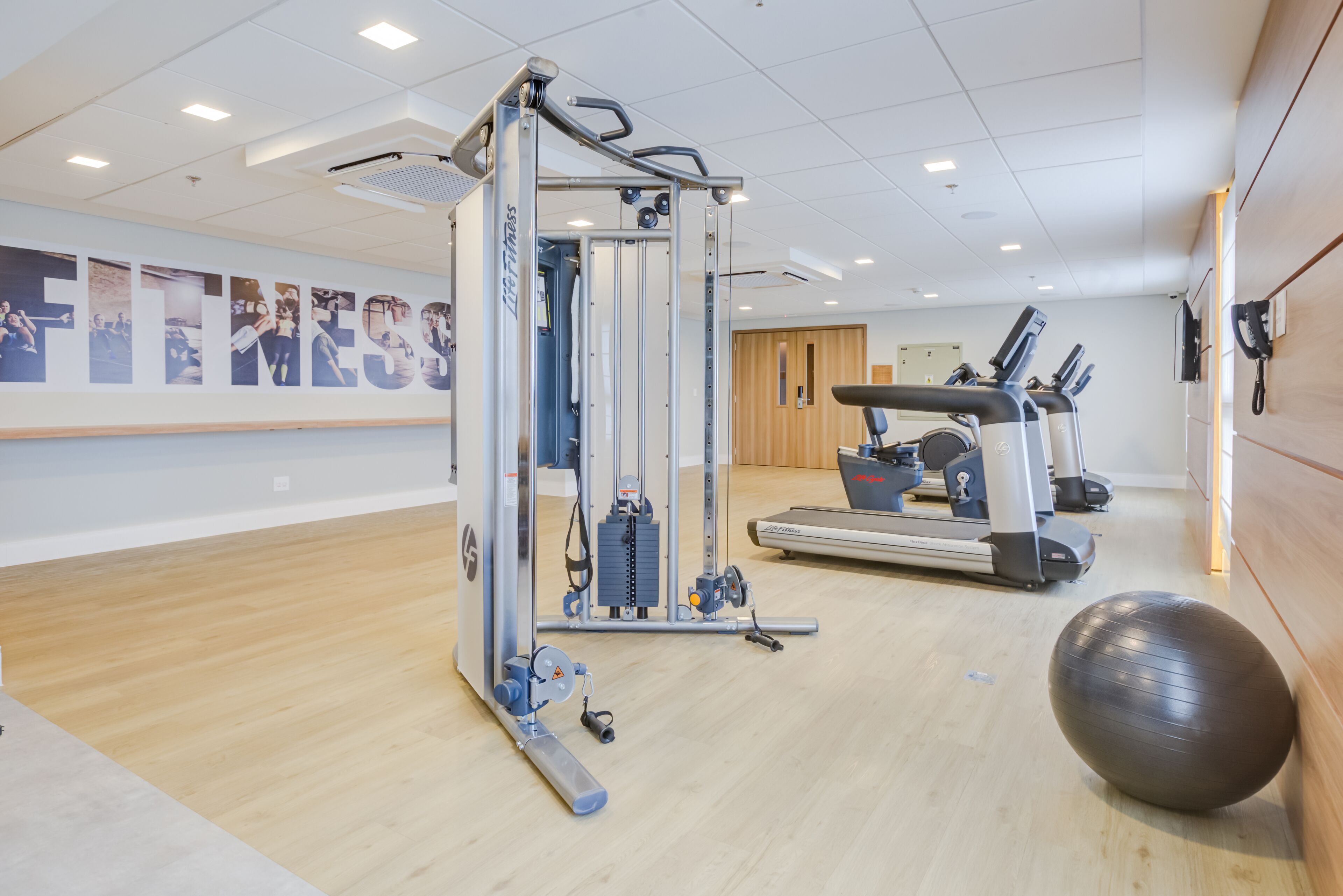 A gym with exercise equipment, a large wall with 'FITNESS' text, and a wooden floor.