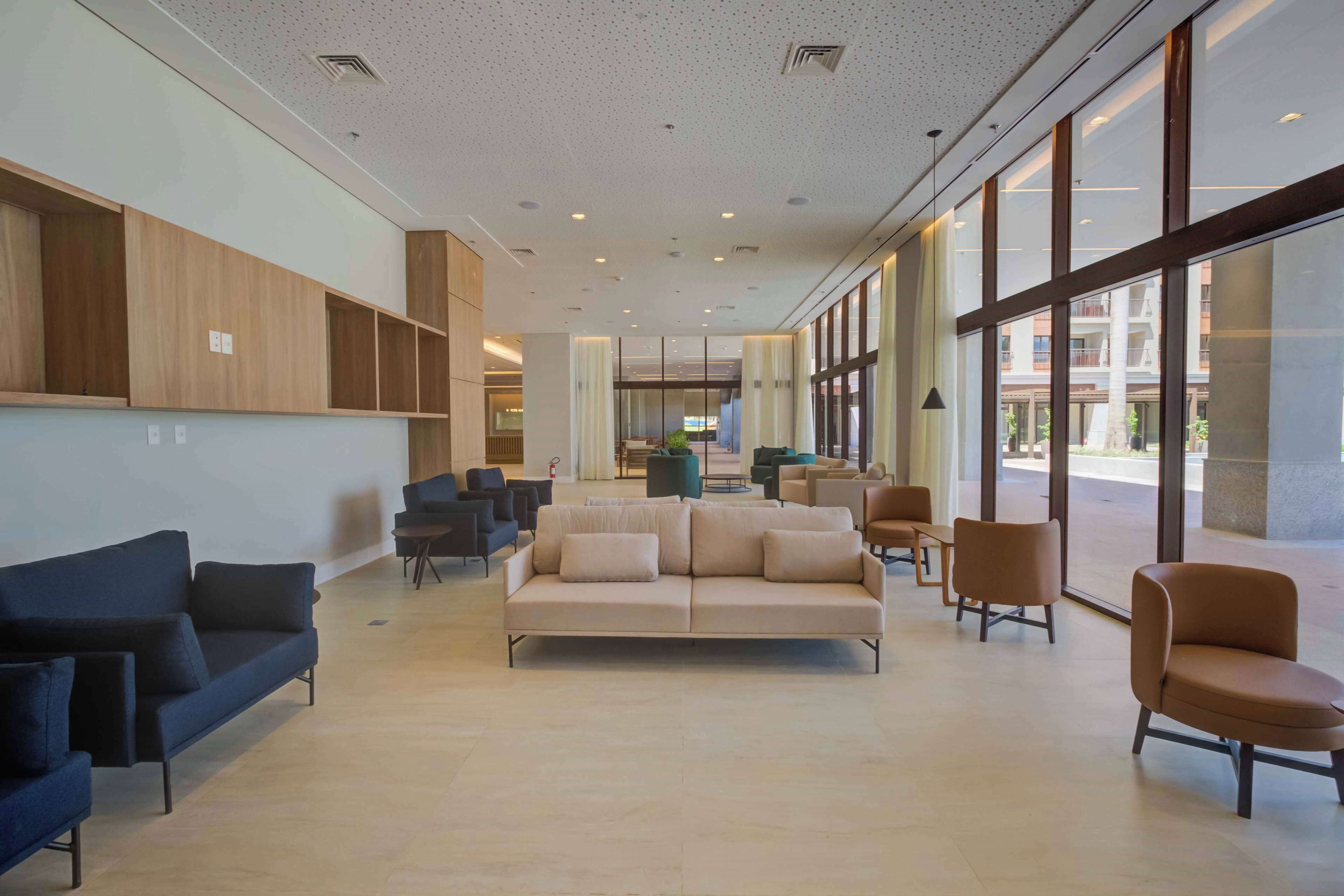 A spacious lobby with a beige sofa, blue and brown chairs, wooden paneling, and large windows offering views of the outside.