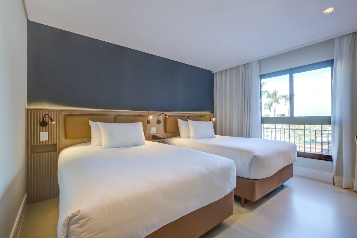 A hotel room with two beds, a large window with curtains, and a view of palm trees.