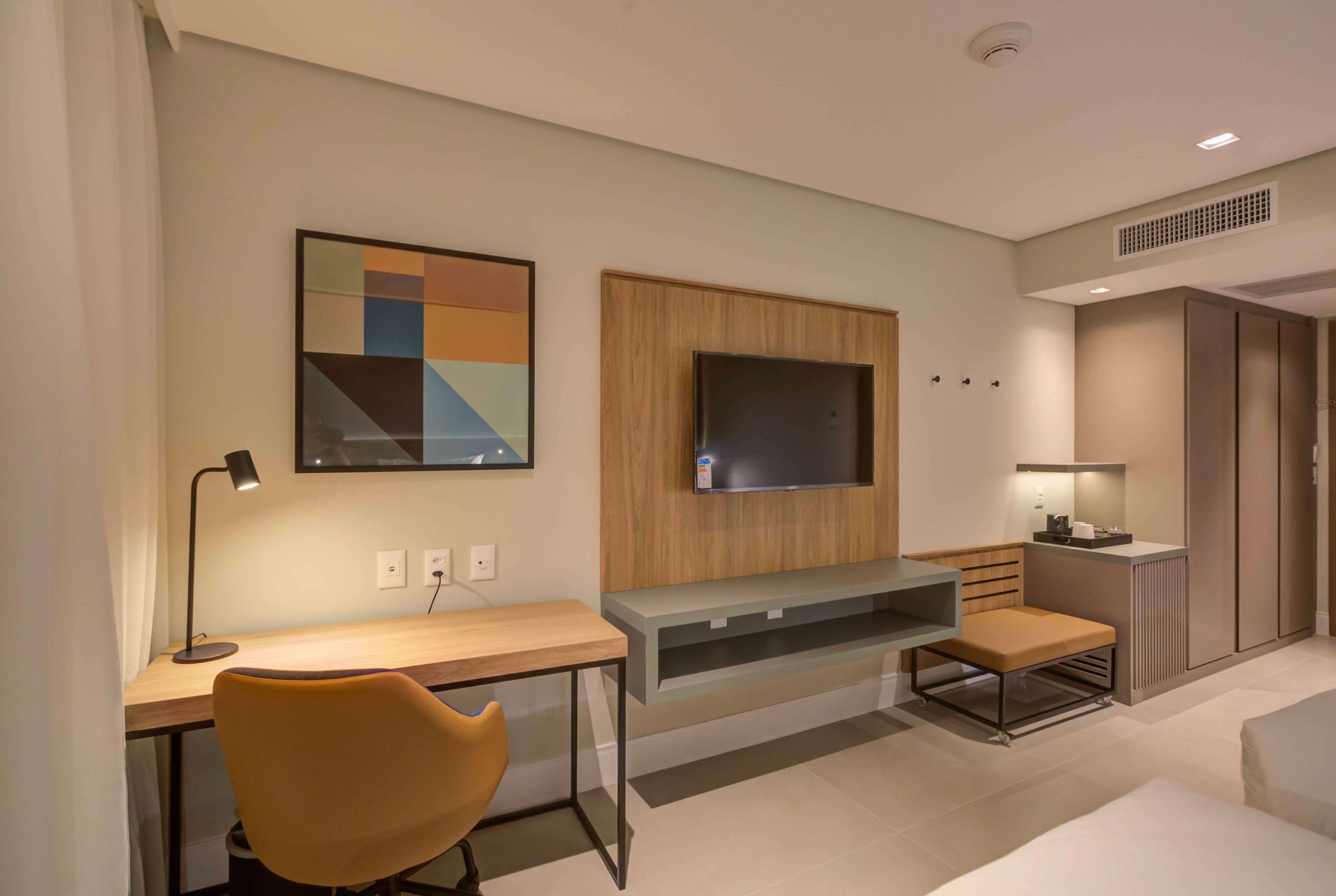 Quarto de hotel moderno com escrivaninha de madeira, uma cadeira confortável, televisão de tela plana na parede e um pequeno espaço de estar com um banco.