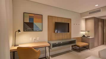 Quarto de hotel moderno com escrivaninha de madeira, uma cadeira confortável, televisão de tela plana na parede e um pequeno espaço de estar com um banco.