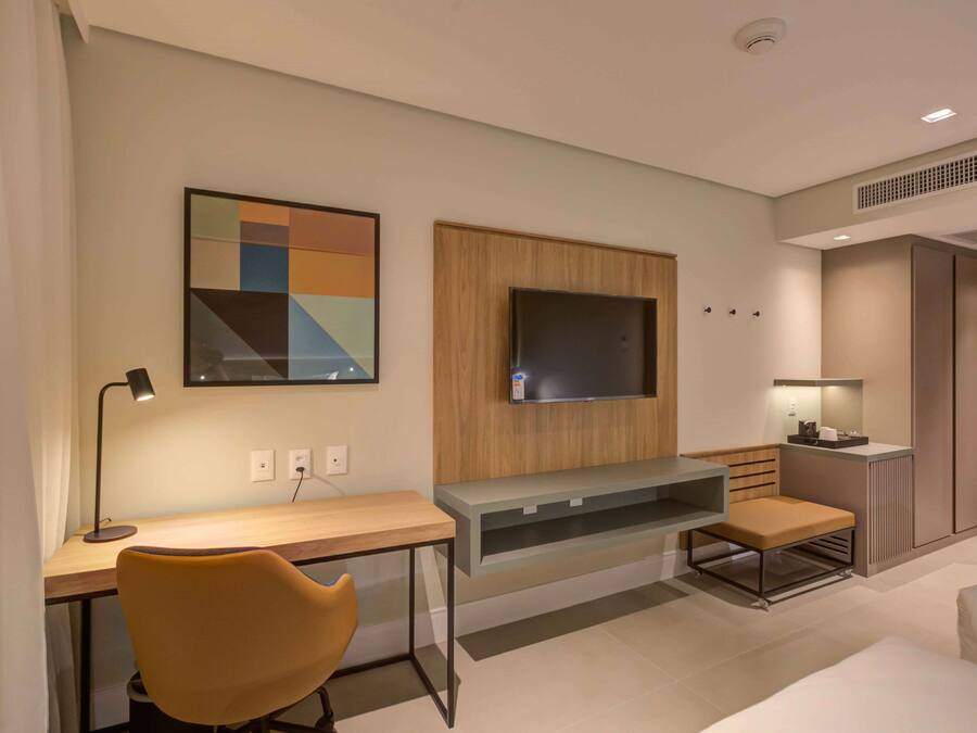 Quarto de hotel moderno com escrivaninha de madeira, uma cadeira confortável, televisão de tela plana na parede e um pequeno espaço de estar com um banco.