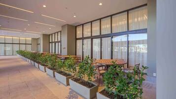 A modern indoor space with rows of tables and chairs, large windows, and potted plants.