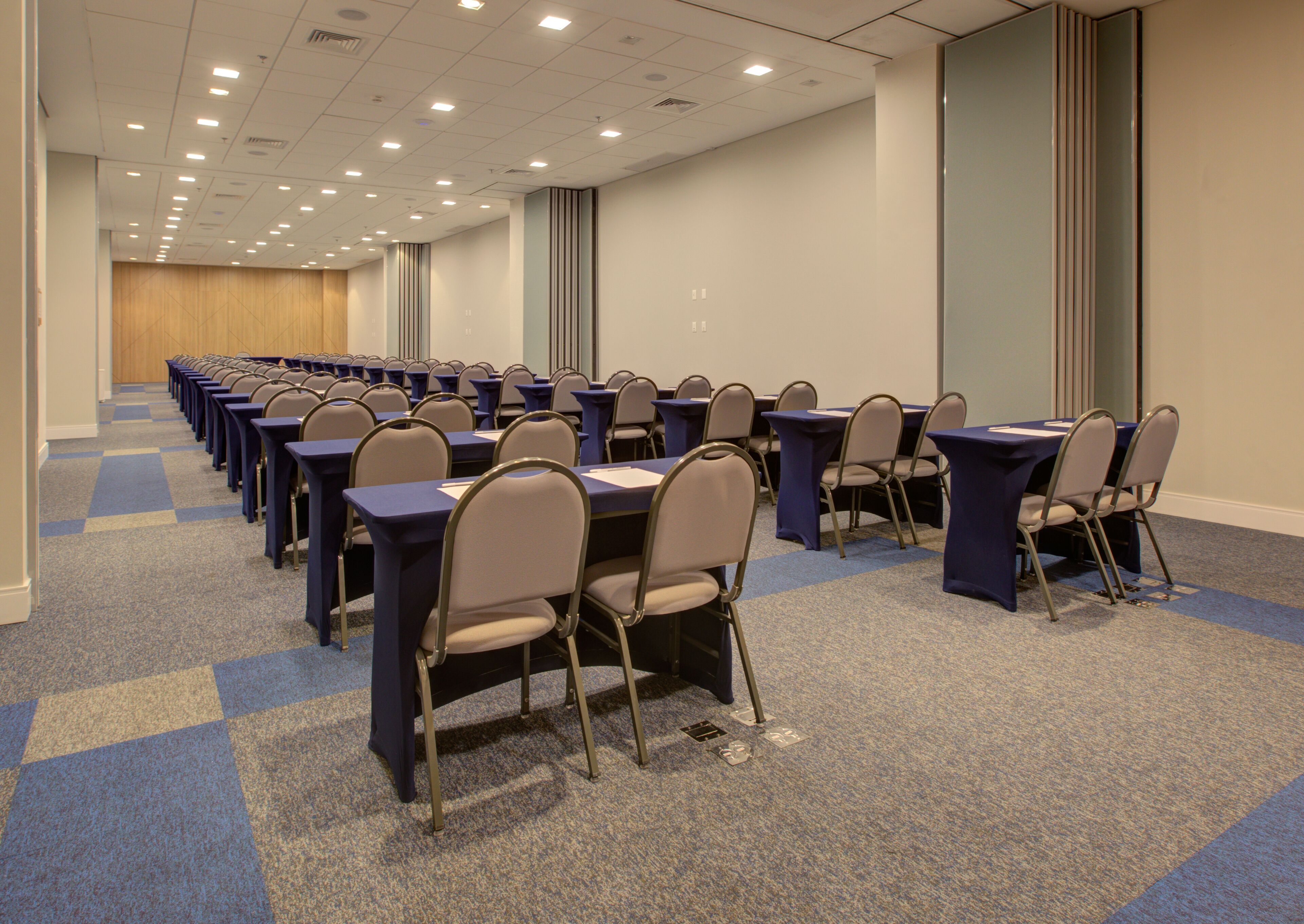 Un salón de conferencias con filas de mesas y sillas, piso alfombrado y un largo pasillo con puertas.
