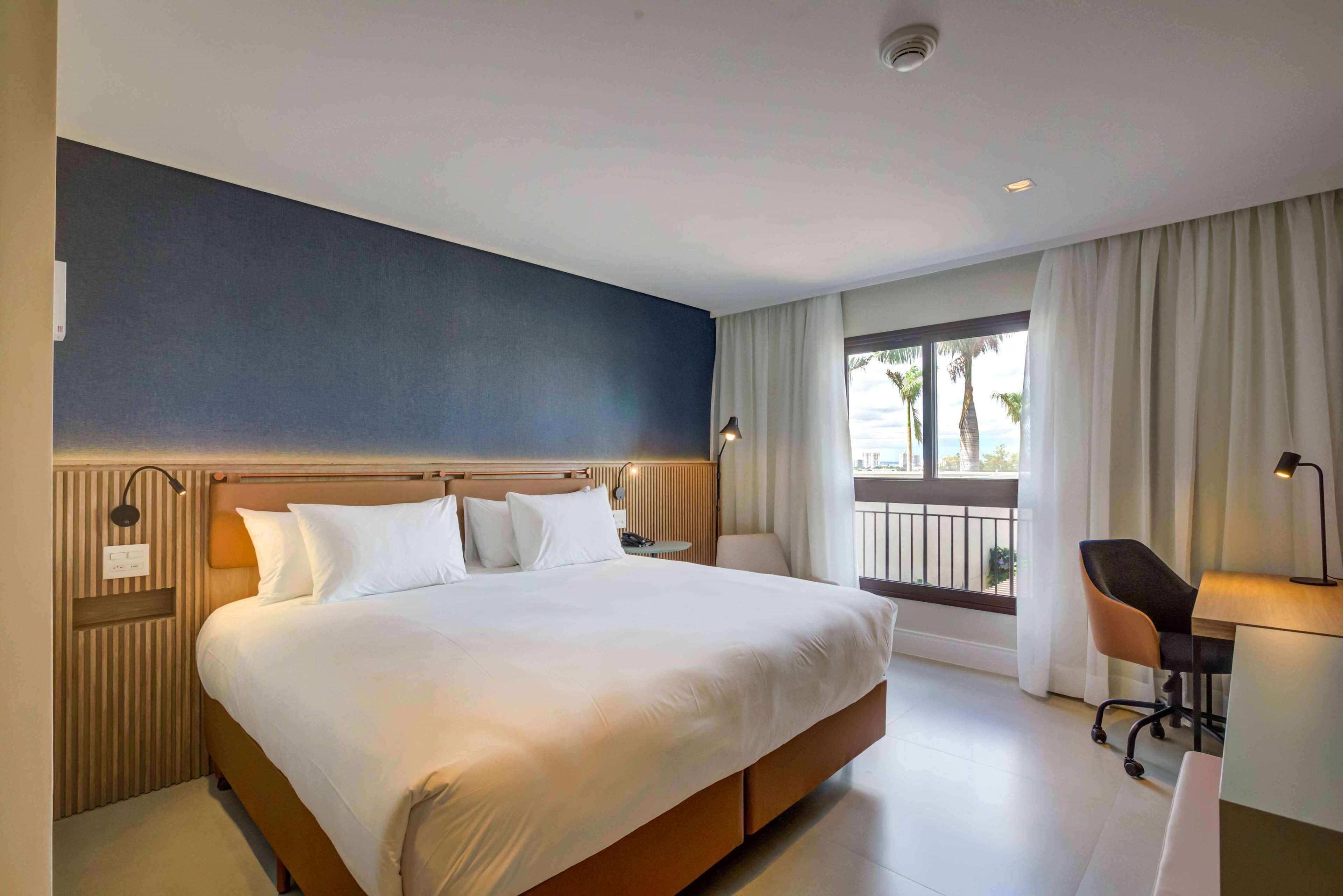 Habitación de hotel con una cama grande, un escritorio con silla, una ventana con cortinas y vistas a palmeras.
