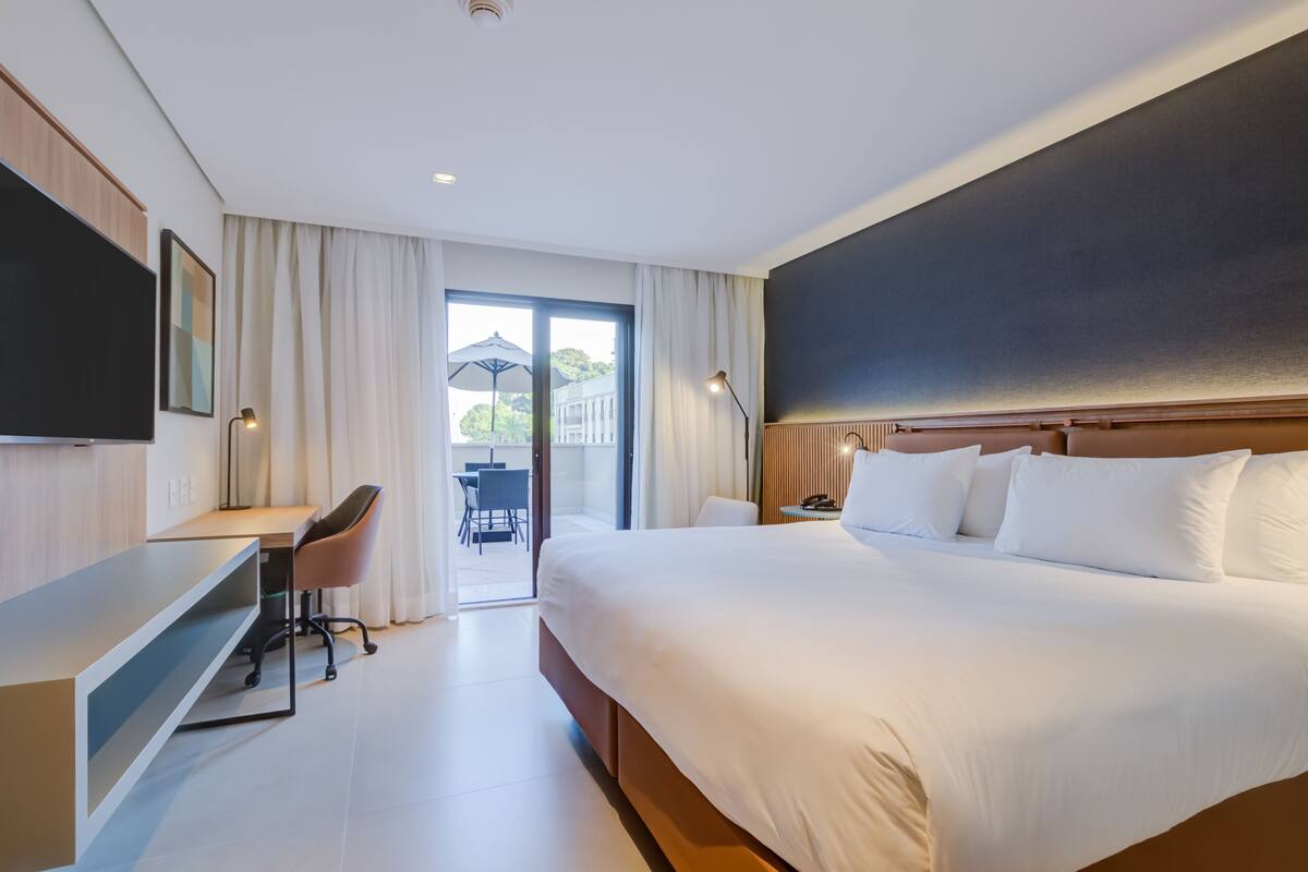A modern hotel room with a large bed, a desk, a chair, a TV, and a balcony with an umbrella.
