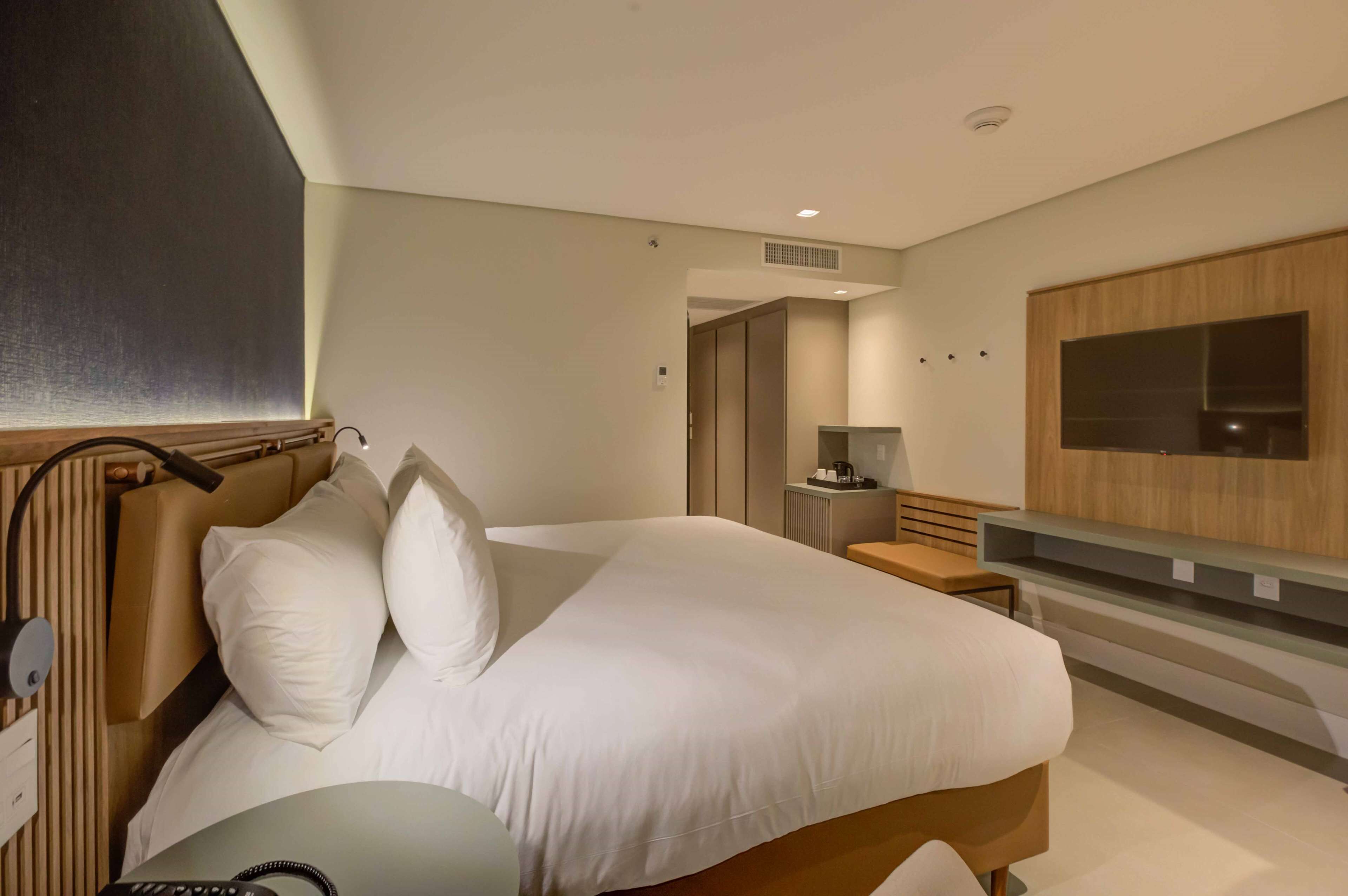 A modern hotel room with a large bed, bedside tables, a TV mounted on the wall, and a small desk area.