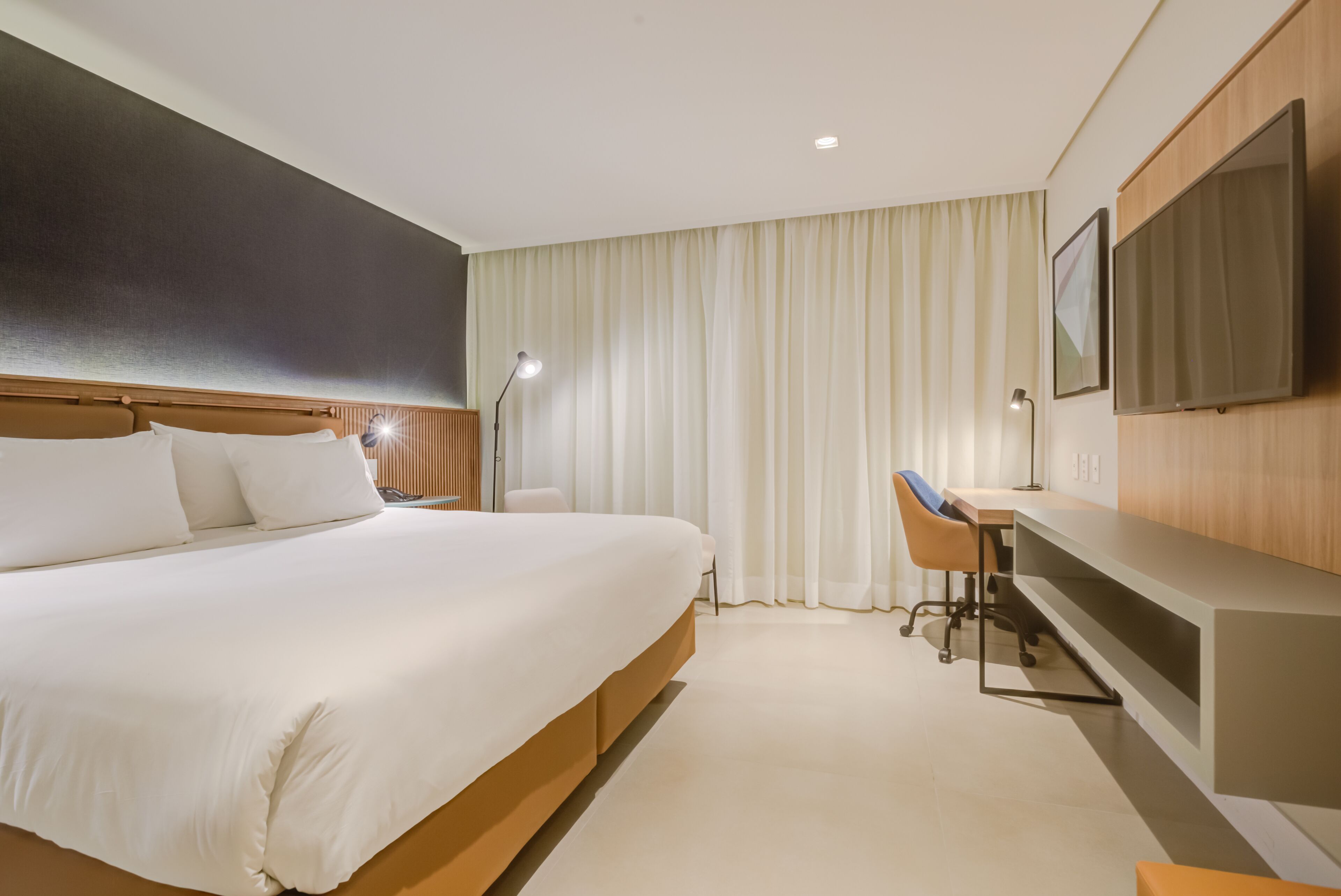 A hotel room with a large bed, a desk, a chair, a TV, and a window with curtains.