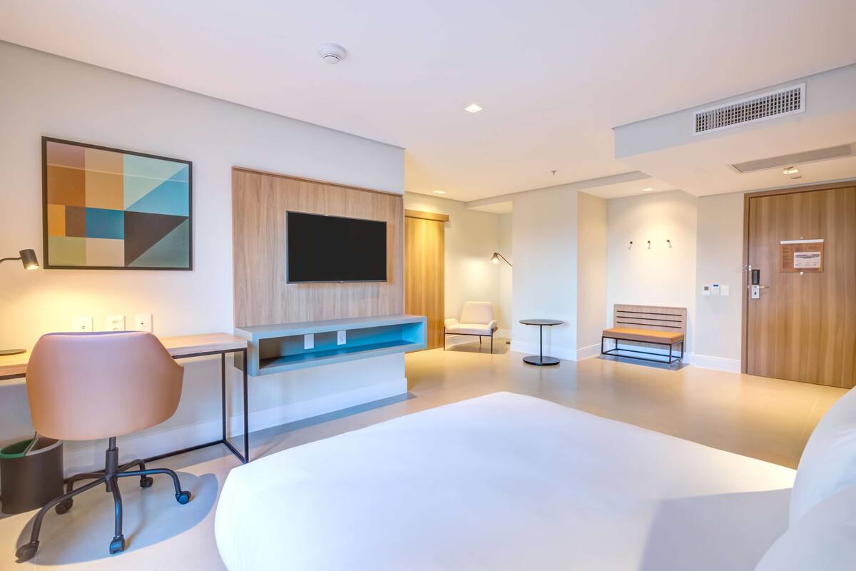 A modern hotel room with a large bed, a desk with a chair, a flat-screen TV, a bench, and a small table.