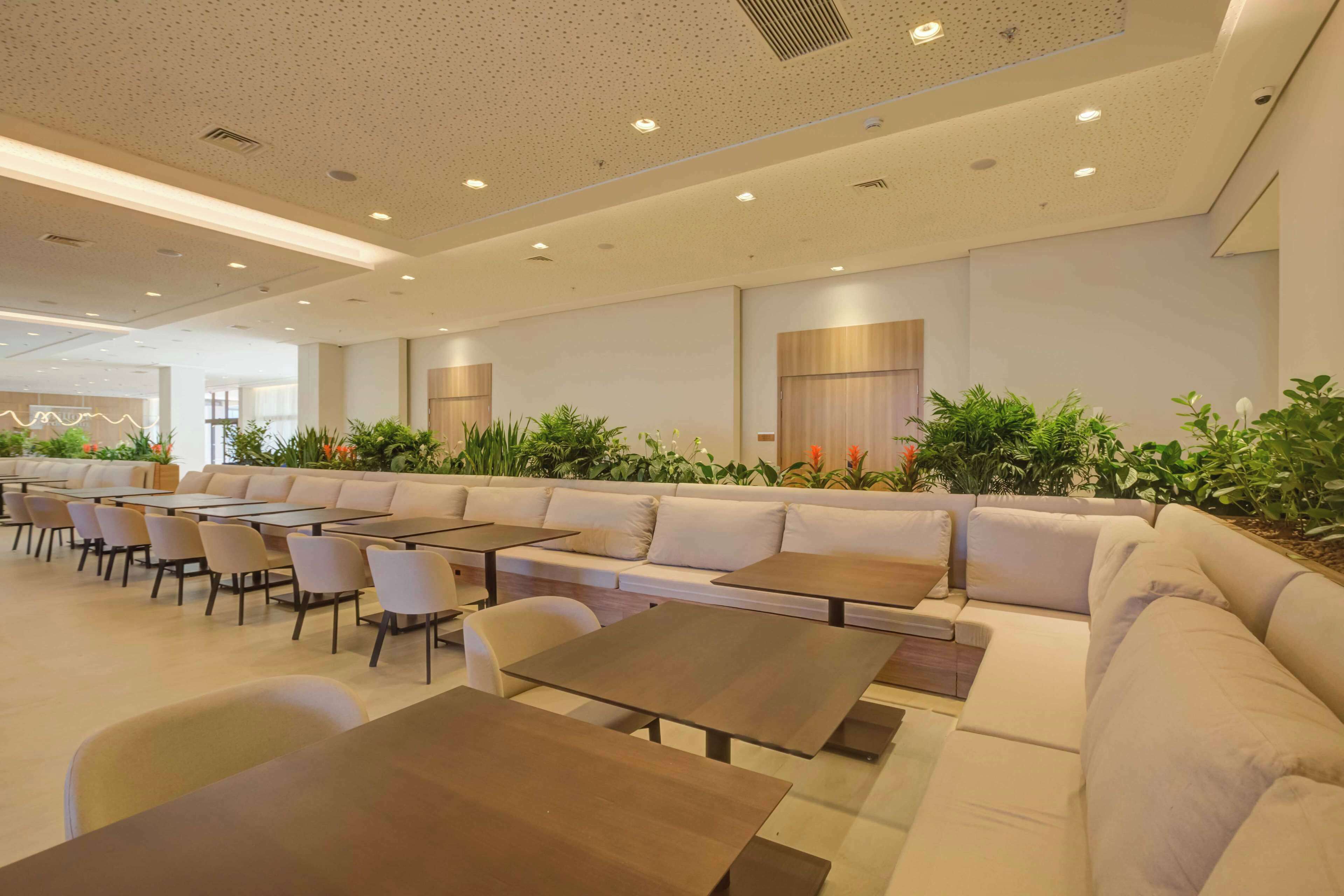 A modern indoor seating area with tables and sofas, surrounded by potted plants.