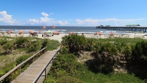 A beach with a wooden boardwalk, beach umbrellas, and a pier in the background. - Lavish Oceanfront Townhome- Whispering Palms (Folly Beach)