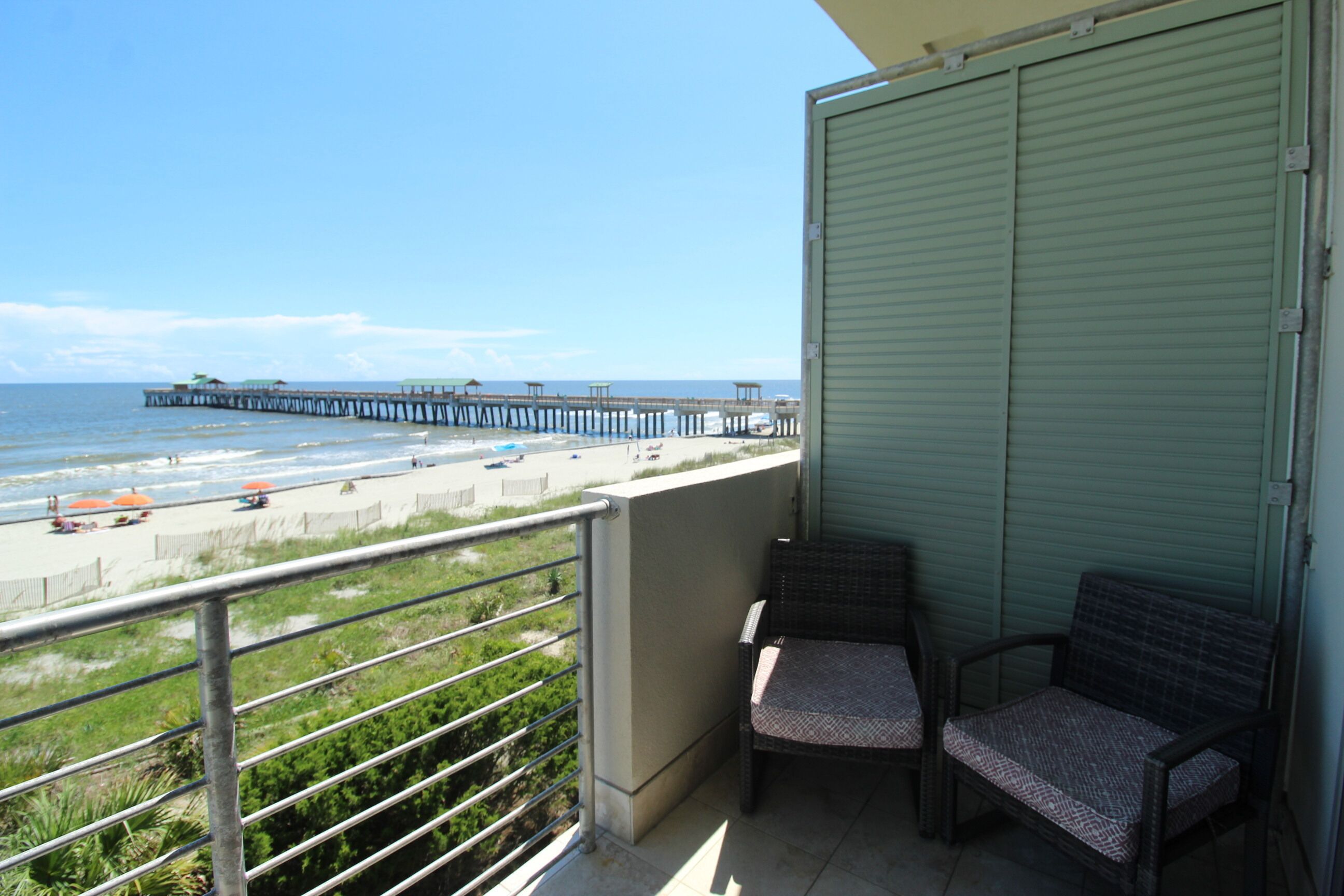 Een balkon met twee rieten stoelen, uitzicht op het strand en een pier op de achtergrond.