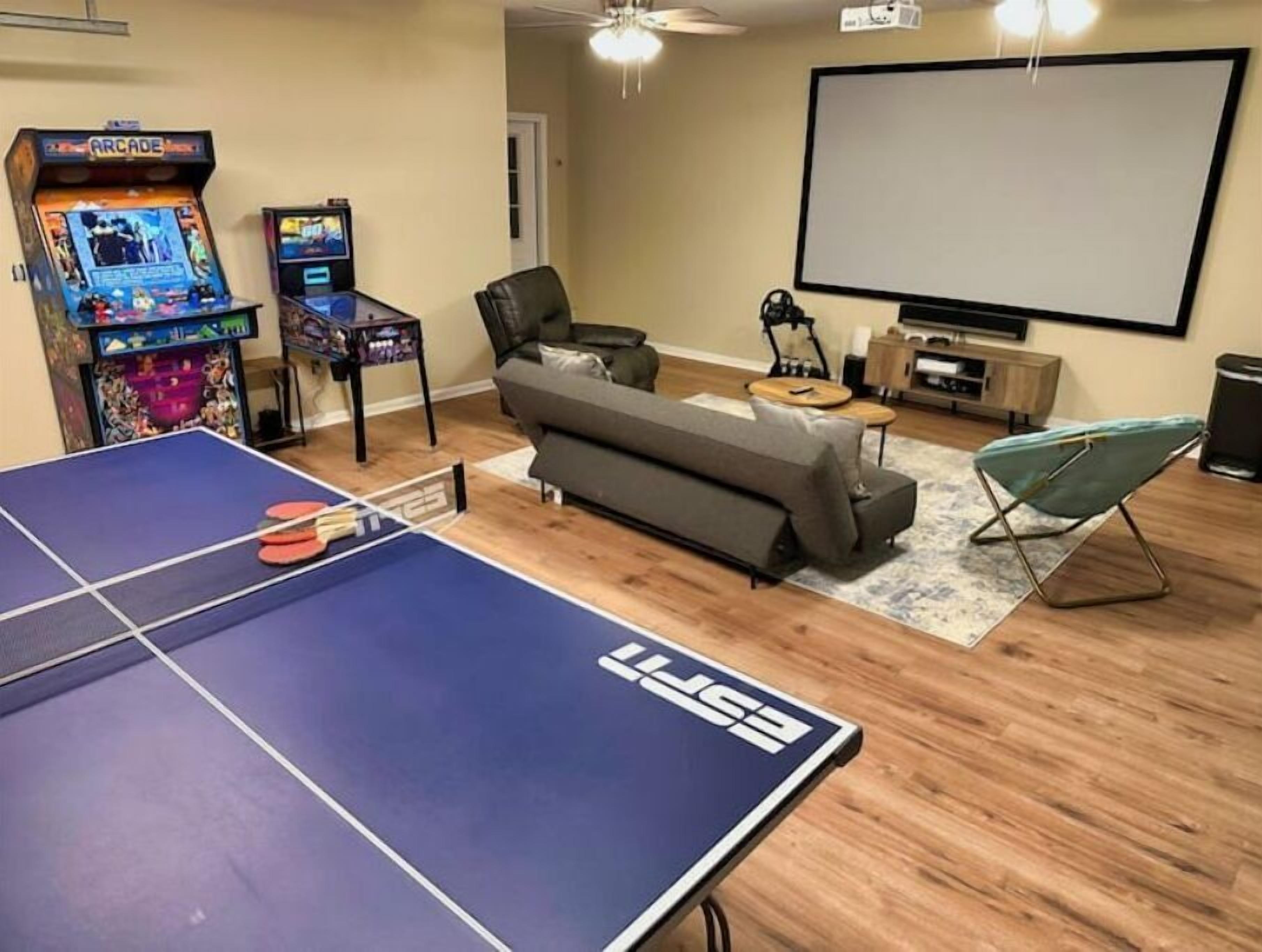 A room with a ping pong table, two arcade machines, a sofa, a projector screen, and a TV.