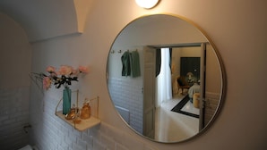 Un baño con un espejo redondo, un estante con un jarrón de flores y una pared de azulejos blancos.