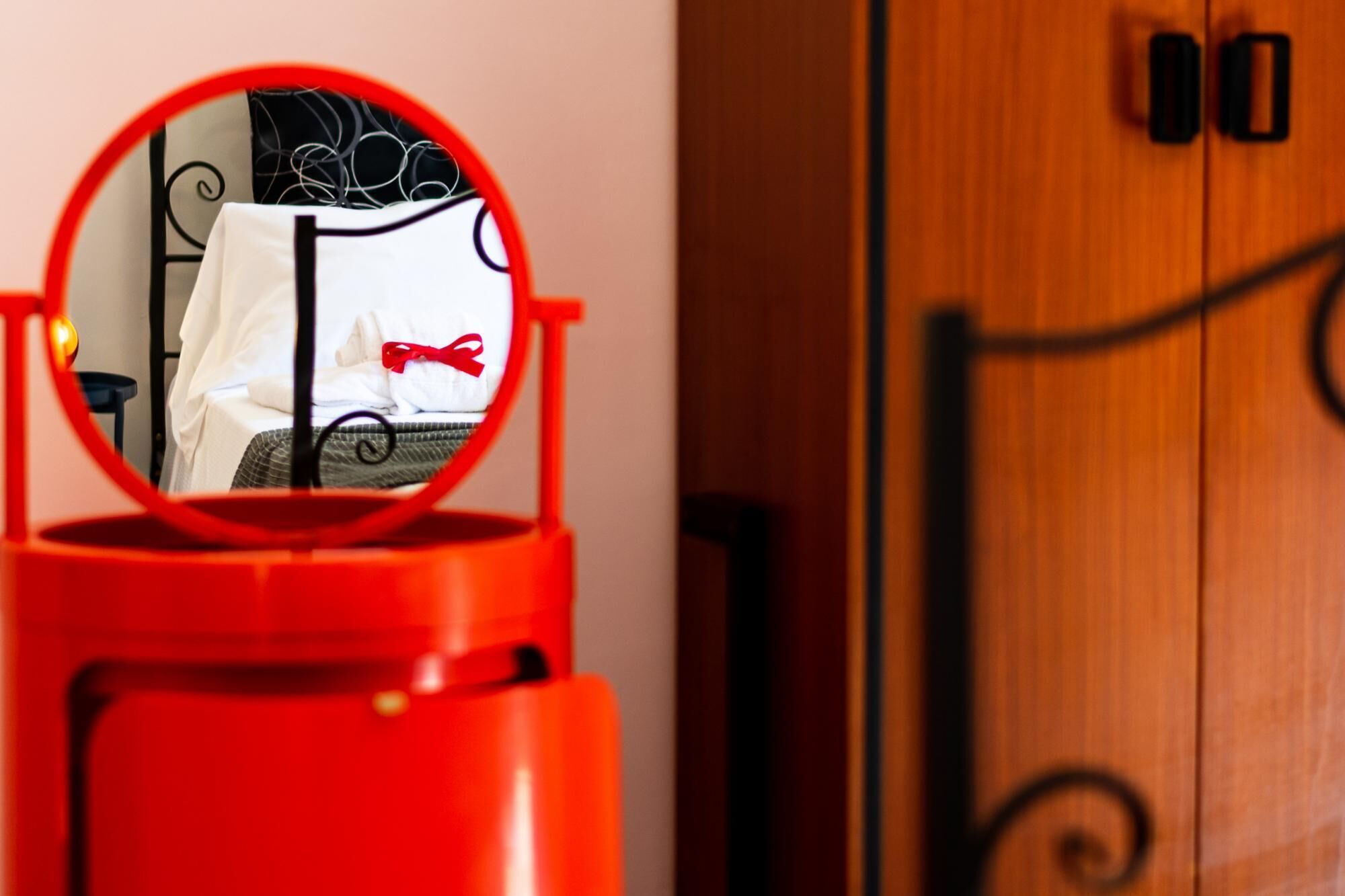 Une poubelle rouge surmontée d’un miroir rond qui reflète un lit avec une literie blanche et un nœud rouge.