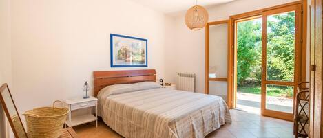 Un dormitori amb un llit, una tauleta de nit, una làmpada, un quadre al mur i vistes a l'exterior a través d'una porta corredissa de vidre.