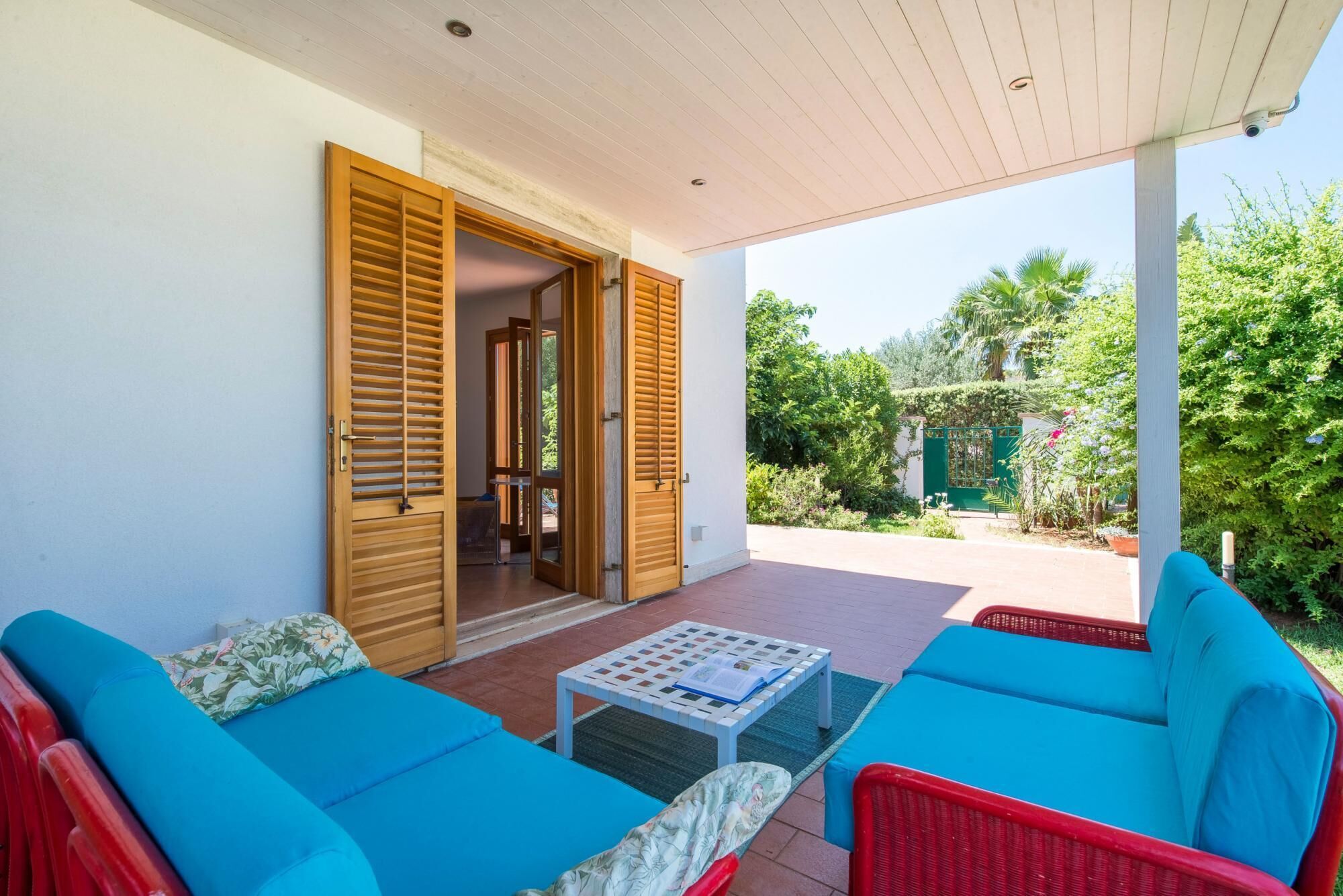 A covered patio with blue and red furniture, a white table, and a wooden door leading to an interior space.