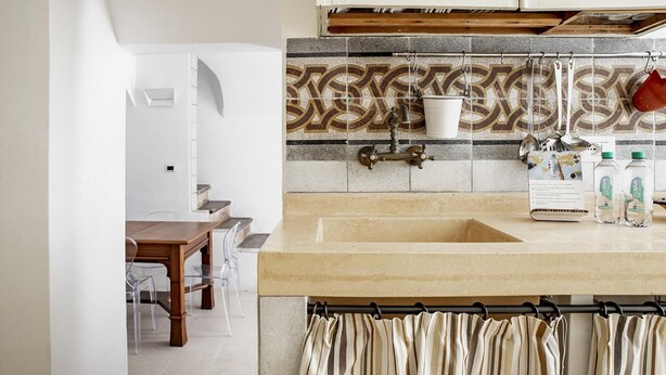 A kitchen with a beige countertop, a sink, and a patterned backsplash. There are clear chairs and a wooden table in the adjacent dining area. - Casa Ibla Duplex con Terrazza by Wonderful Italy (Ragusa)