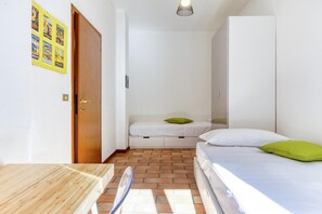 Um quarto pequeno e iluminado com uma mesa de madeira, uma cama de solteiro com almofadas verdes e um guarda-roupa.