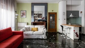 Ein Wohnzimmer mit einem roten Sofa, einem weißen Sofa mit goldenem Kissen, einem Bücherregal und einer Küchenzeile mit einer Theke und einer Hängeleuchte.