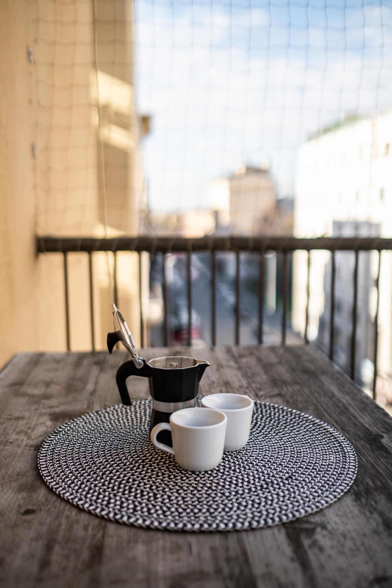 A wooden table with a black and white woven placemat, a coffee pot, and two white mugs on a balcony with a metal railing.