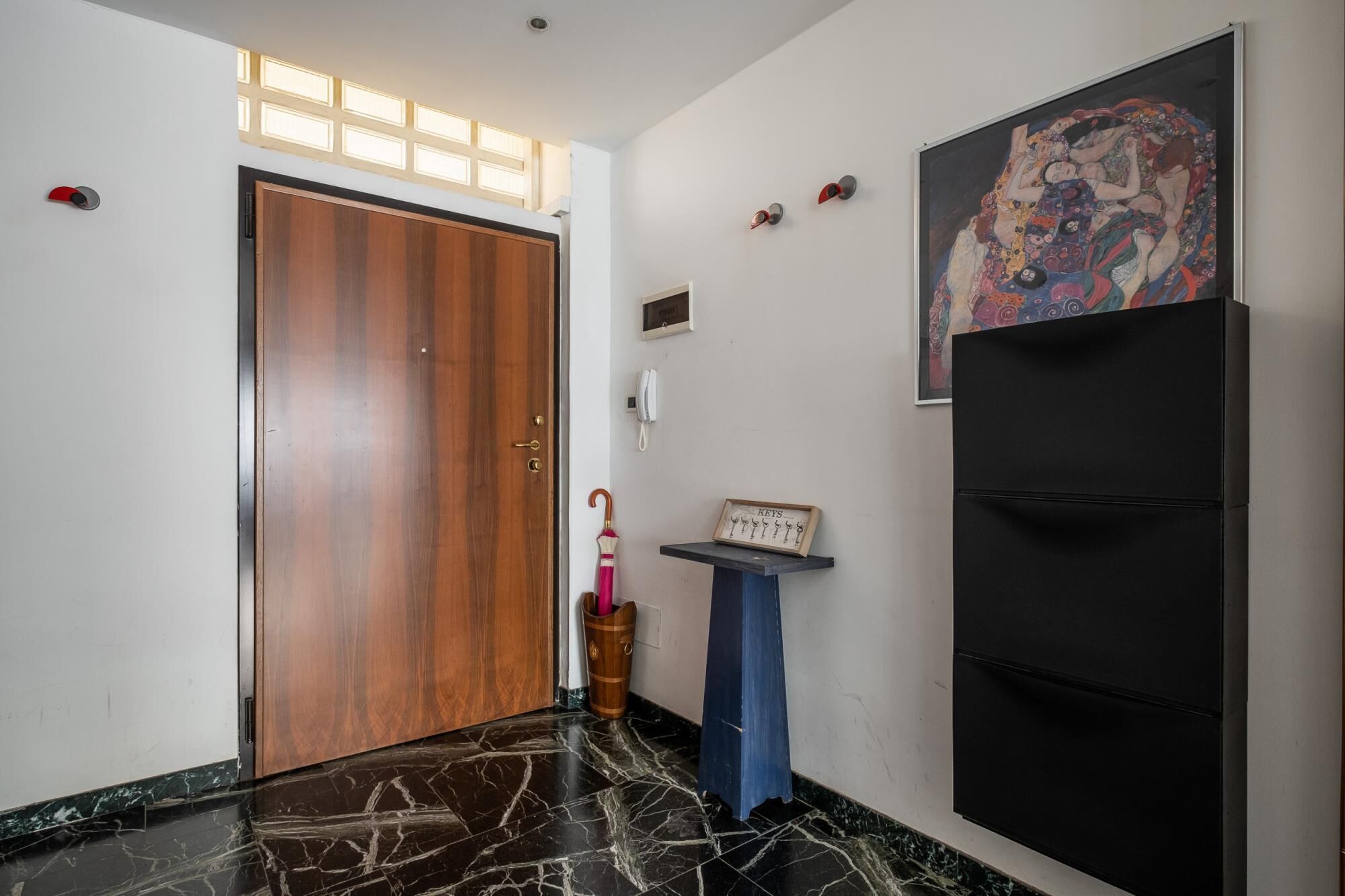Un pasillo con una puerta de madera, un refrigerador negro, una pequeña mesa con un letrero que dice 'KEYS' y un cuadro colorido en la pared.
