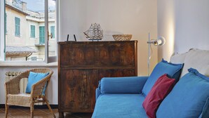 A room with a blue sofa, wicker chair, wooden cabinet, and a view of buildings through a window.