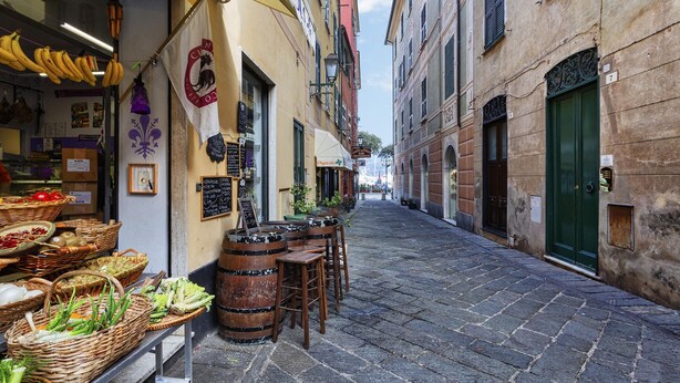 A narrow cobblestone street with a café on the left, displaying fresh produce and wooden barrels.
