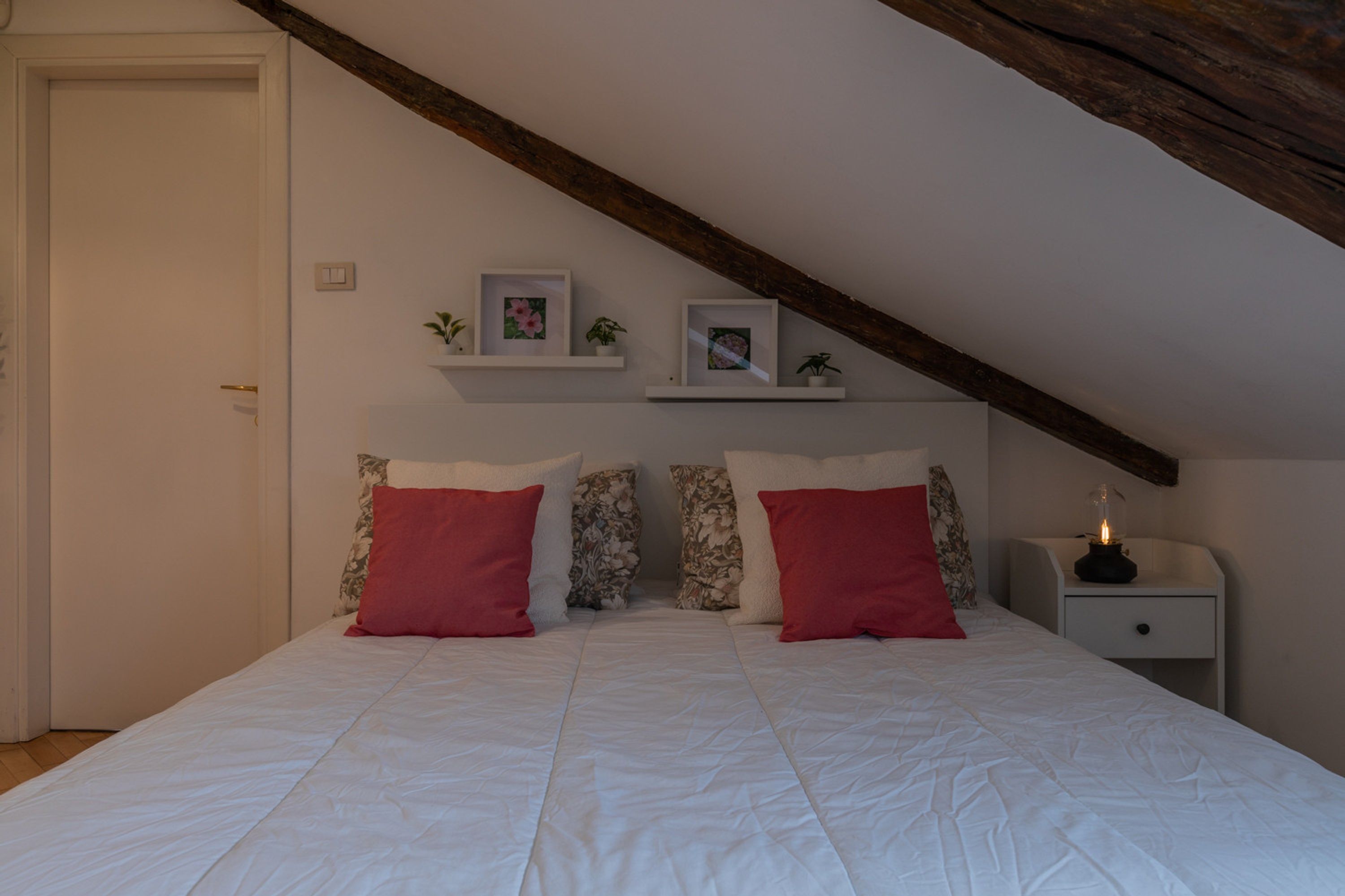 Un dormitorio con techo inclinado, una cama con ropa blanca y almohadas rojas, una mesita de noche con lámpara y estantes en la pared con cuadros y plantas.