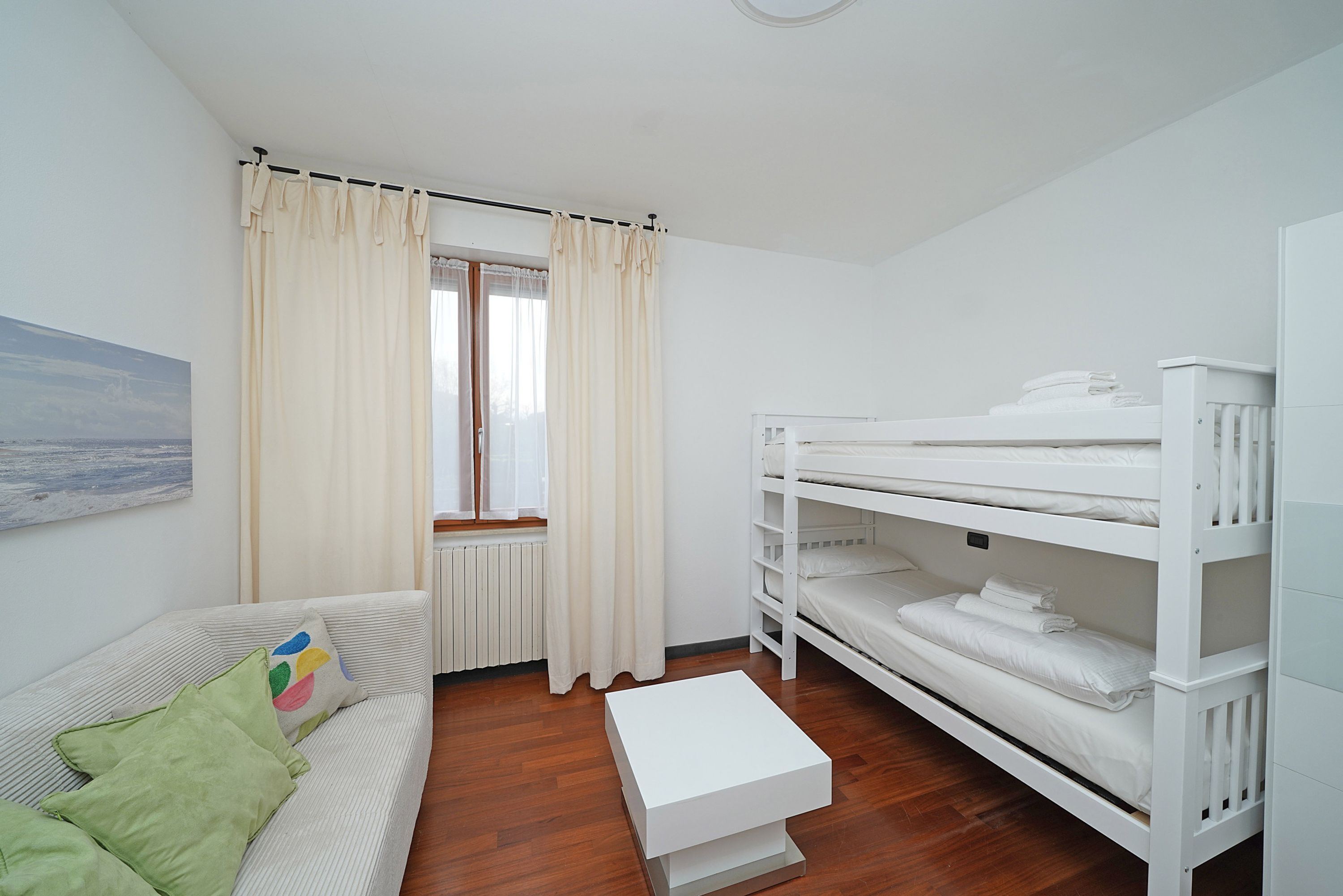 A room with a bunk bed, a sofa with green cushions, a white coffee table, and a window with curtains.