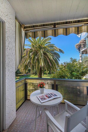 Ein Balkon mit einem weißen Stuhl, einem kleinen runden Tisch mit einer Zeitschrift und Blick auf eine Palme und Grünpflanzen.