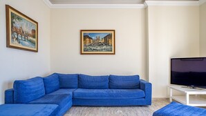 A living room with a blue sectional sofa, a white TV stand, and two paintings on the wall. - Ortigia Seafront Apartment by Wonderful Italy (Syracuse)