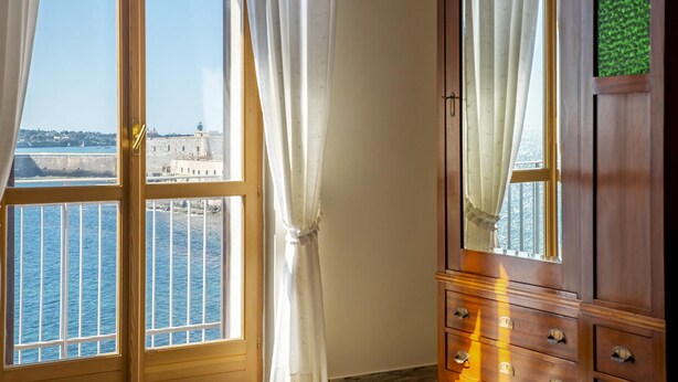 A room with a large window overlooking a coastal area, a wooden dresser with a mirror, and a view of a historic building across the water. - Ortigia Seafront Apartment by Wonderful Italy (Syracuse)