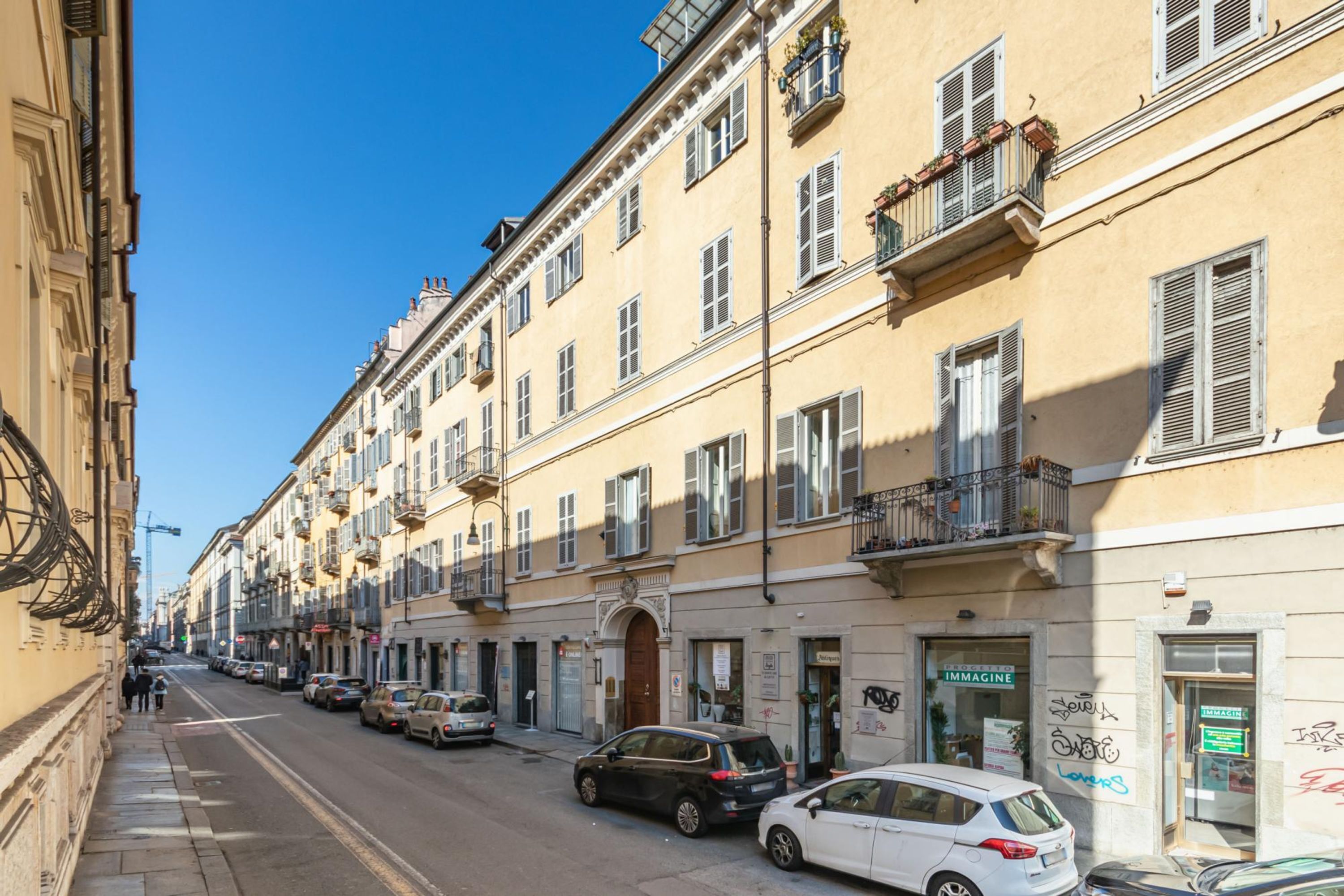 A street with parked cars and buildings with balconies and shutters.