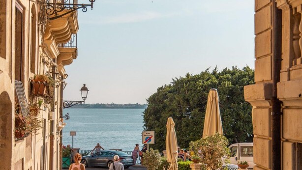A cobblestone street with outdoor seating, a body of water in the background, and buildings with classical architecture. - Appartamento di Stile a Ortigia by Wonderful Italy (Syracuse)