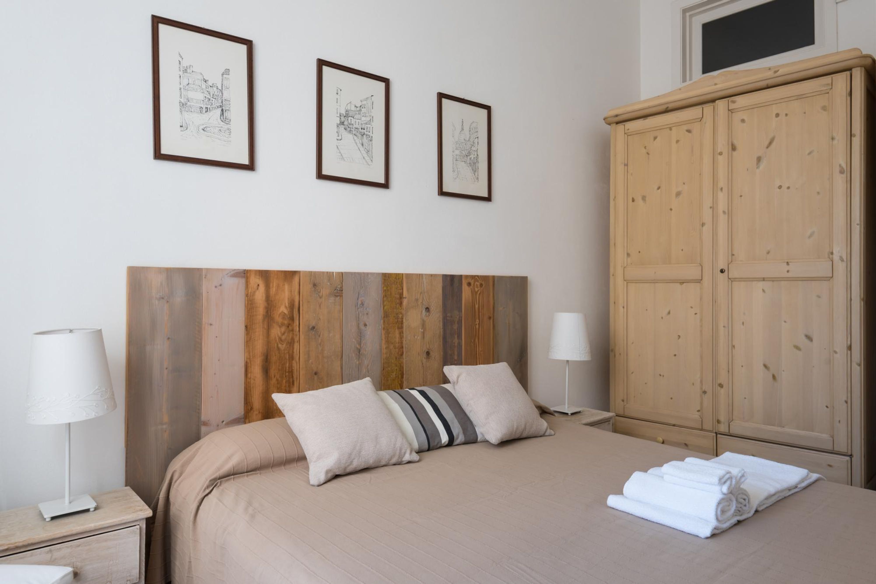 Un dormitorio con cabecera de madera, cama con ropa de cama color beige, dos almohadas, una mesita de noche con una lámpara y un armario.
