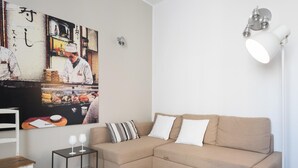 A living room with a beige sectional sofa, a small side table with two wine glasses, a striped rug, and a wall-mounted lamp. - Appartamento di Stile a Ortigia by Wonderful Italy (Syracuse)