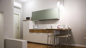 Et lille køkken med hvide skabe, en mikrobølgeovn og et spiseområde med metalstole.