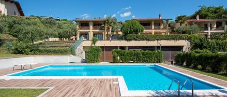 A swimming pool with a wooden deck, surrounded by lush greenery and a multi-story building with balconies.