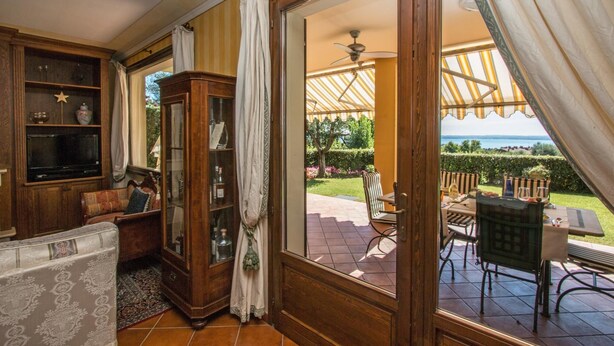 A room with a wooden cabinet, a sofa, and a view of an outdoor patio with a table and chairs. - Le Ortensie 1 Apartment by Wonderful Italy (Soiano del Lago)