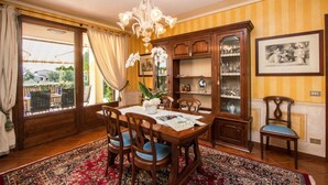 A dining room with a wooden table, chairs, a chandelier, a cabinet with glass doors, and a large window with curtains. - Le Ortensie 1 Apartment by Wonderful Italy (Soiano del Lago)