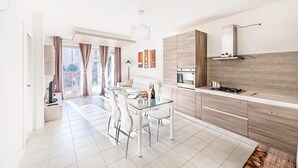 A modern kitchen with a built-in oven and gas stove, a dining area with a glass table and white chairs, and a view of the city through large windows. - Le Corti Caterina B2 Apartment by Wonderful Italy (Lonato del Garda)