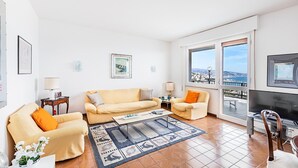 A spacious living room with a large sofa, a smaller armchair, a coffee table, and a TV. There are two side tables with lamps, and a large window offering a view of the city. - Le Orchidee 19 by Wonderful Italy (Sanremo)