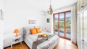 A bedroom with a bed, a bedside table with a lamp, a tray with breakfast, and a large window with a view of greenery. - Le Orchidee 19 by Wonderful Italy (Sanremo)