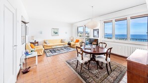 A spacious living area with a sofa, coffee table, and dining table. Large windows offer a view of the sea. - Le Orchidee 19 by Wonderful Italy (Sanremo)
