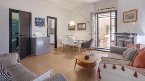A modern living room with a sofa, a coffee table, and a dining area with chairs and a table. There is a kitchen visible through an open door.