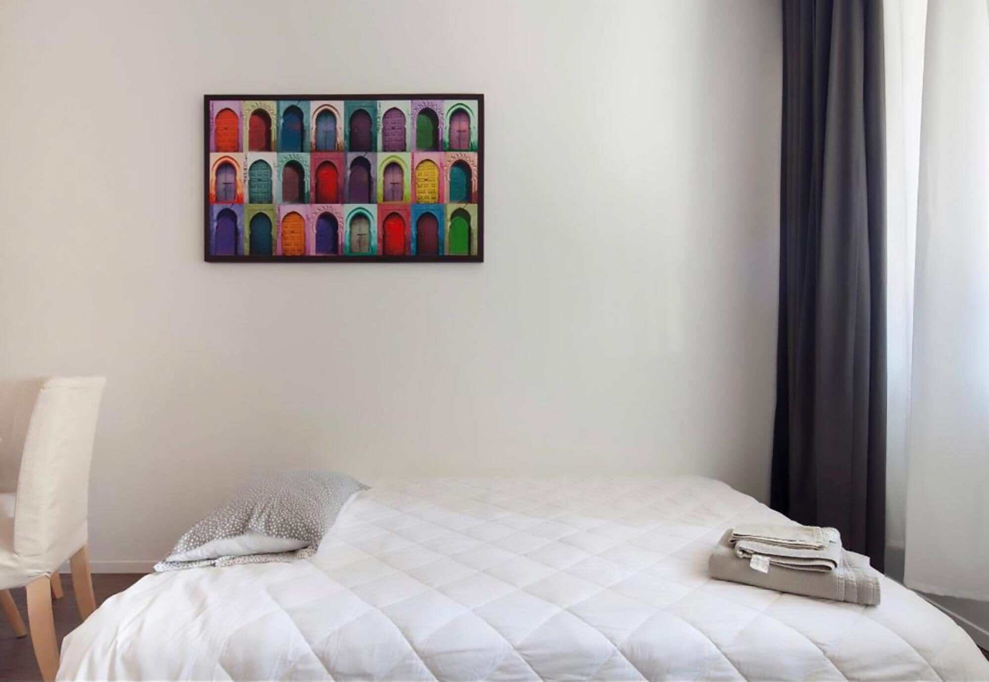 Una habitación de hotel con una cama, una silla, una obra de arte colorida en la pared y una ventana con cortinas.