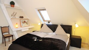 A bedroom with a sloped ceiling, a bed with a dark blanket, two bedside lamps, a small table, a chair, and a shelf with decorative items. - Eckhorst Farm, Ruhwinkel (Güby)