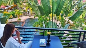 A woman sitting at a table with a book and a cup, overlooking a pool with people swimming. - The Garden Park by Lucky Courtyard (Siem Reap)