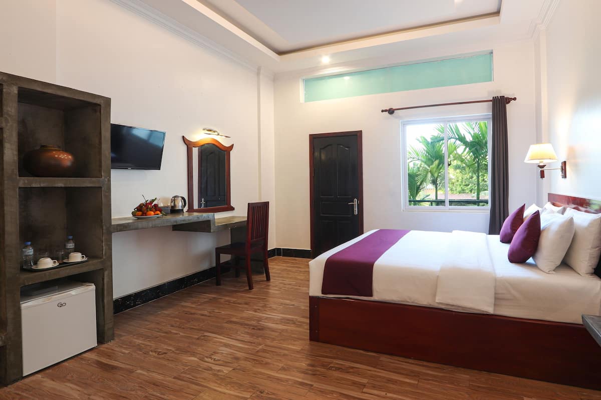 A hotel room with a wooden floor, a bed with white and purple bedding, a desk with a mirror, a TV, and a window with a view of palm trees.